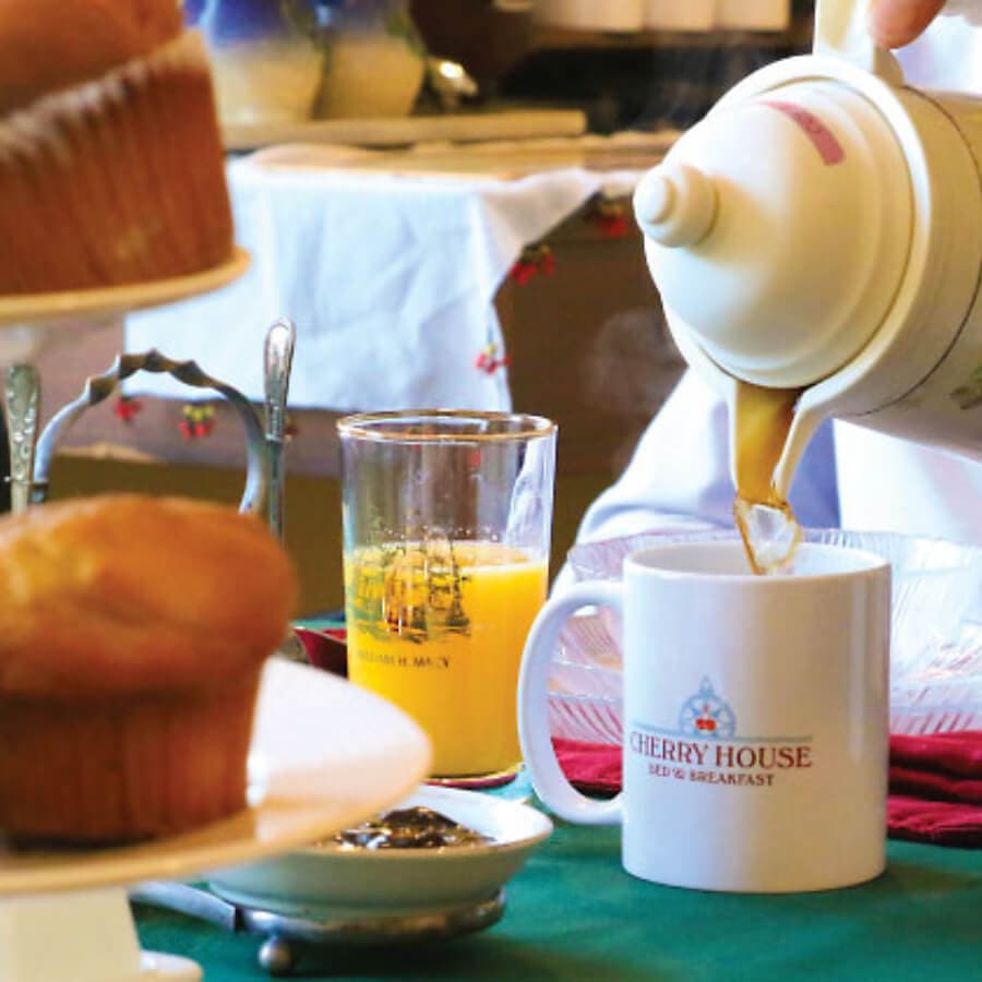 A hand pours coffee into a "Cherry House" mug on a table set with muffins and juice.