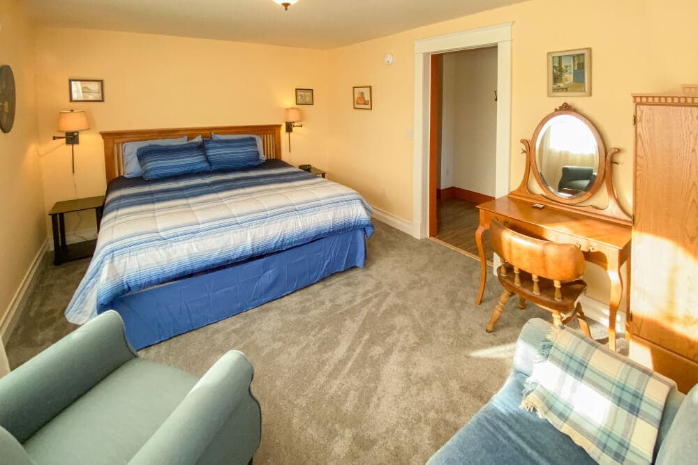 A cozy bedroom featuring a large bed, a vanity with a mirror, and two armchairs in a warm, inviting color scheme.