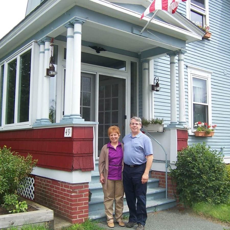 A man and woman stand smiling on the porch of a blue house with an American flag.