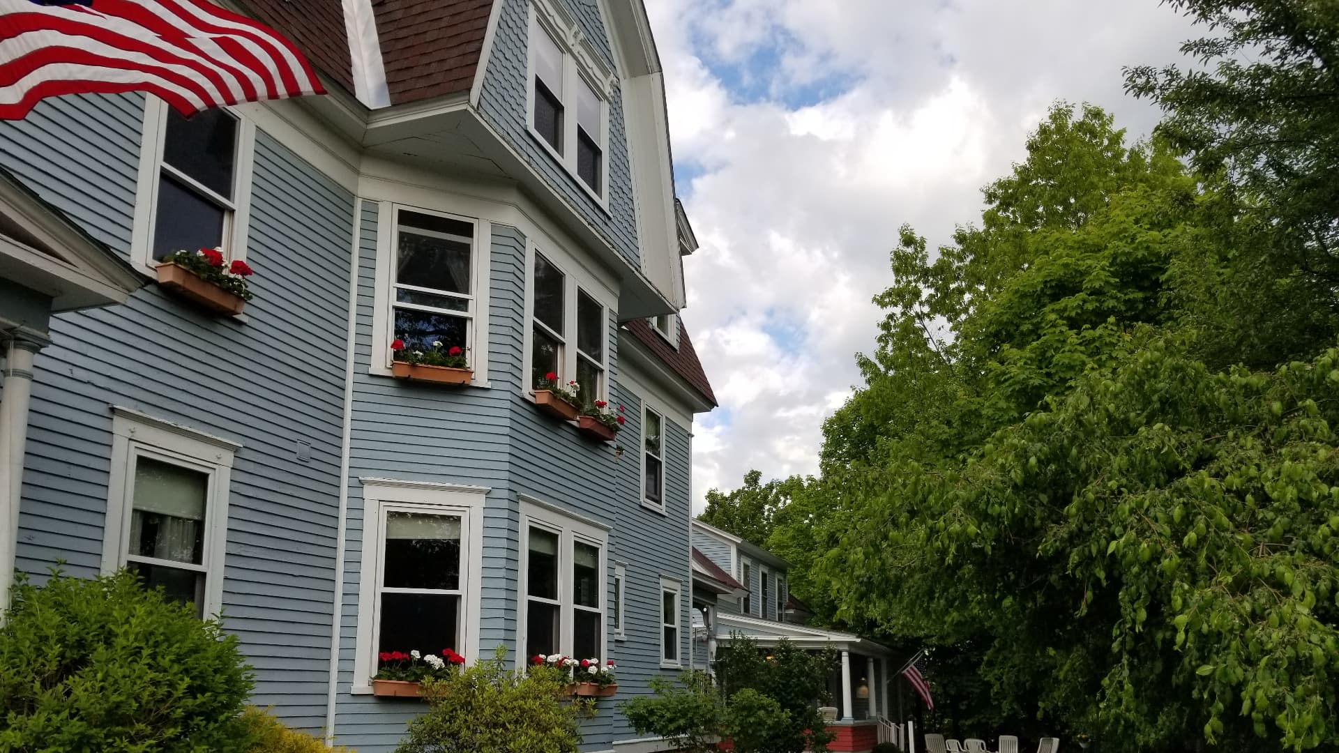 A blue two-story house features flower boxes and an American flag, surrounded by greenery under a cloudy sky.